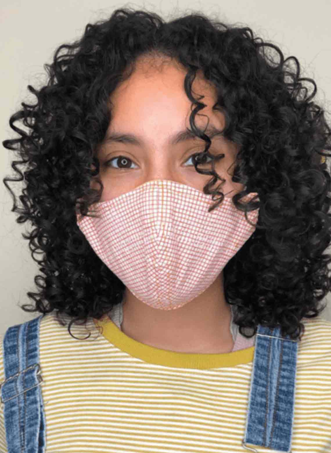 person wearing yellow striped shirt, denim overalls, plaid mask, and shoulder length black curly hair