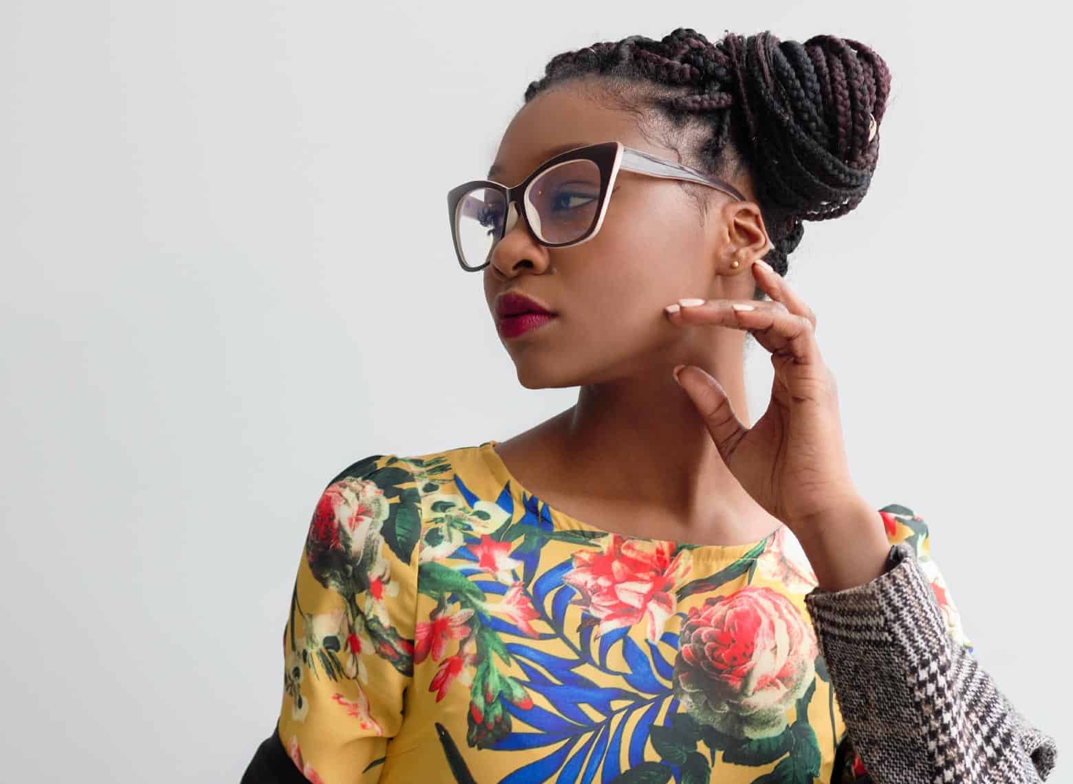 person looking to the side and wearing box braids in a side chignon, also wearing a colorful floral top, glasses, and stud earrings
