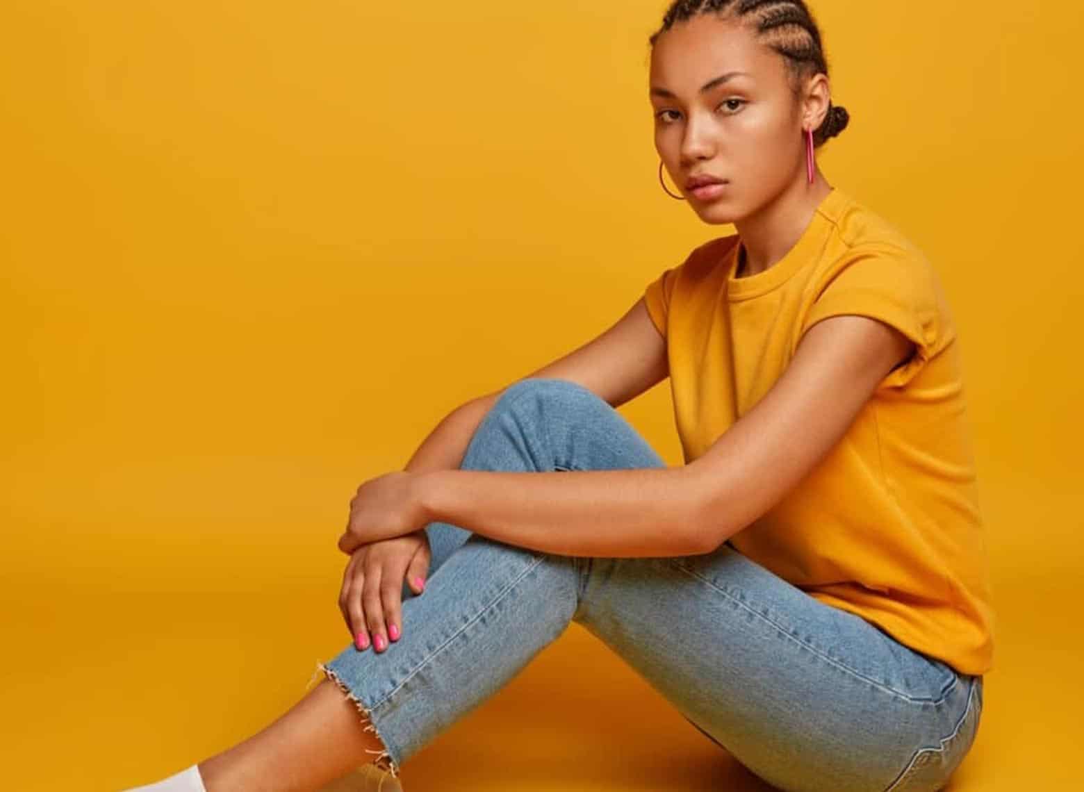 person sitting down in orange room, wearing orange t-shirt, blue jeans, white socks and cornrows