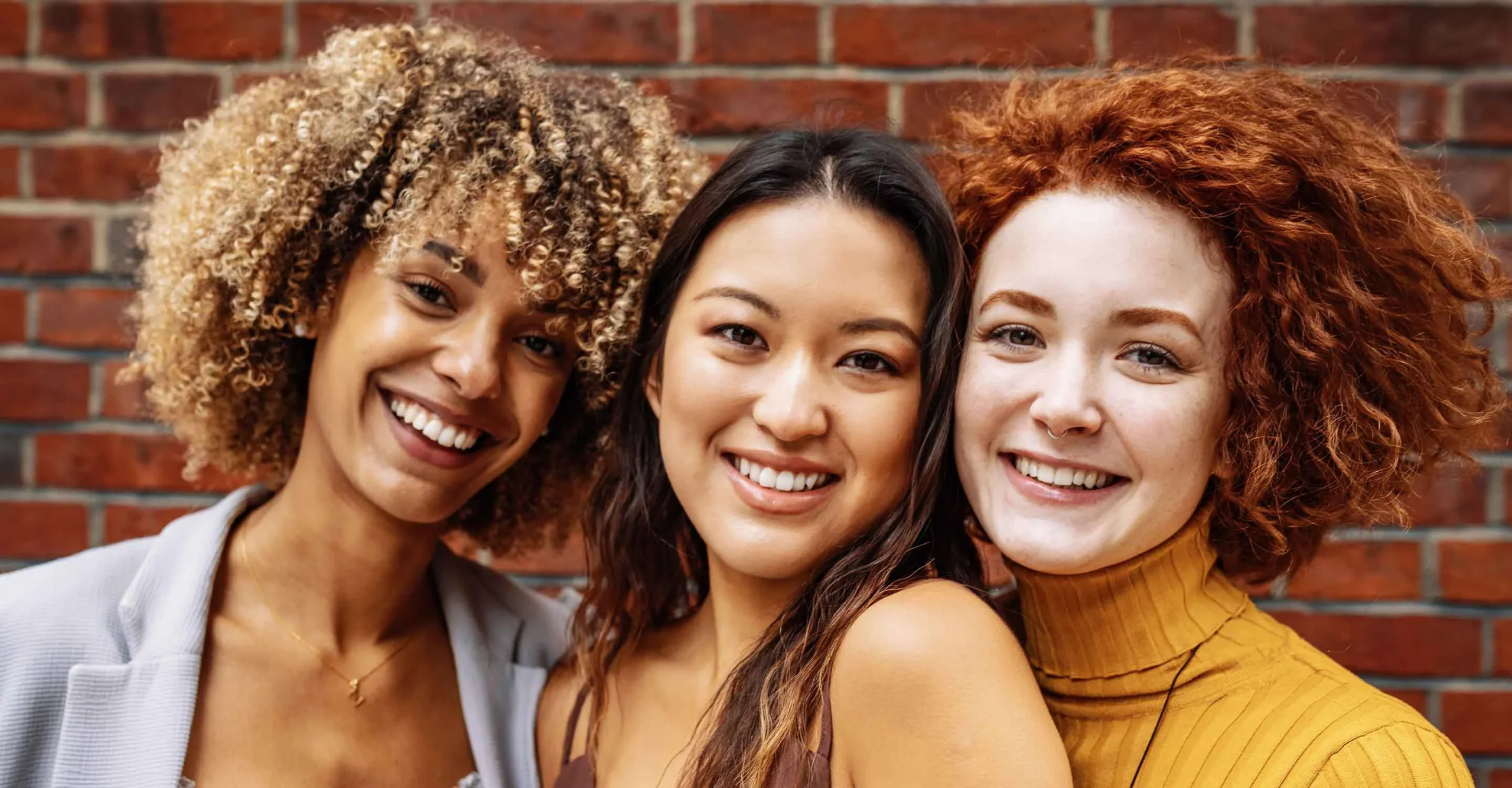 Three women with different types of curly and wavy hair smile together.