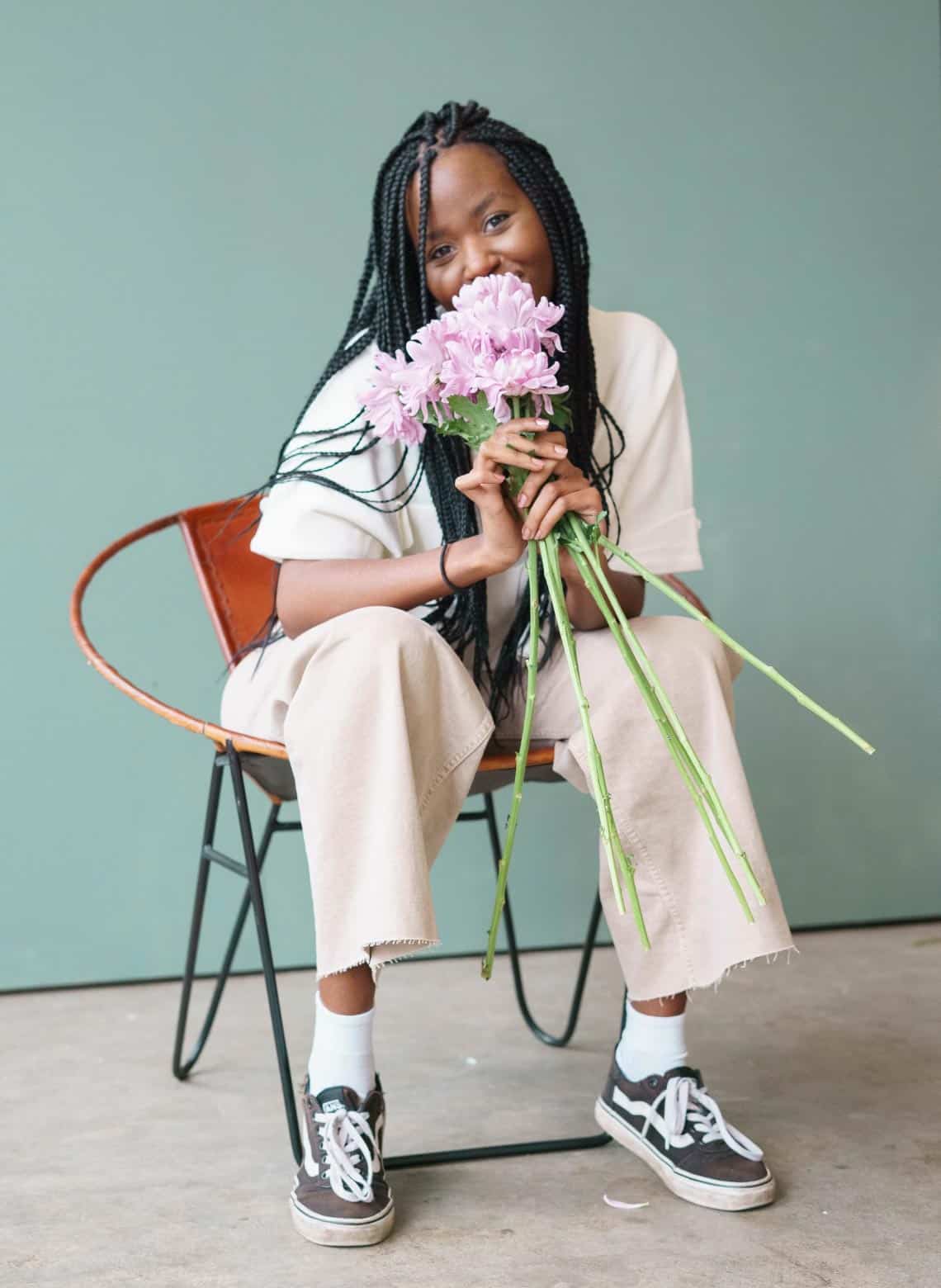 person sitting in a chair holding pink flowers with long stems, wearing baggy khaki shirt and pants, with long knotless braids