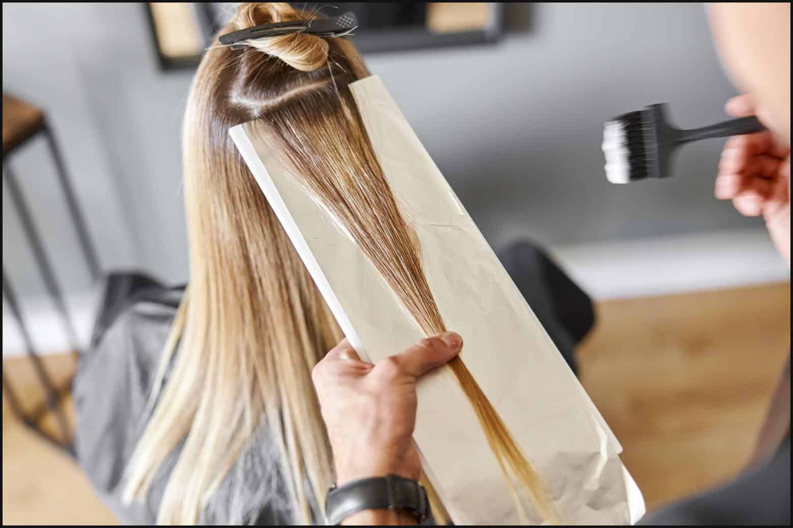 close up on person’s dark blonde hair while stylist applies bleach to a section of their hair