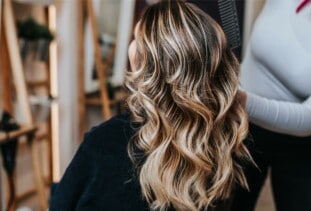 person with brunette hair and blonde balayage highlights sitting in salon chair facing the mirror while stylist stands and works on hair