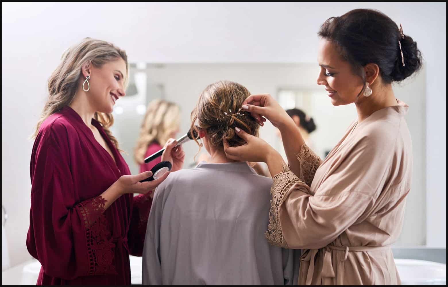 Back view of makeup artist wearing a magenta robe and hair stylist wearing a beige robe working on client