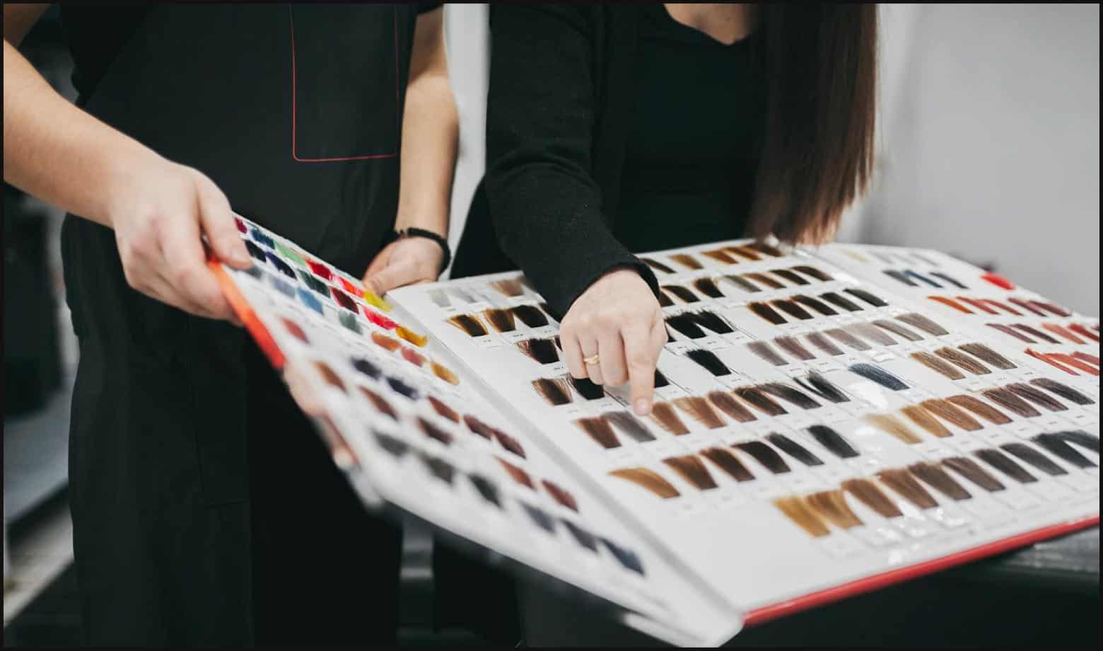 client and stylist, both dressed in black, looking through a book of hair color samples in a salon