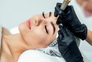 A woman is wrapped in a towel and wearing a hair net while she lays still getting her eyebrows tattooed.
