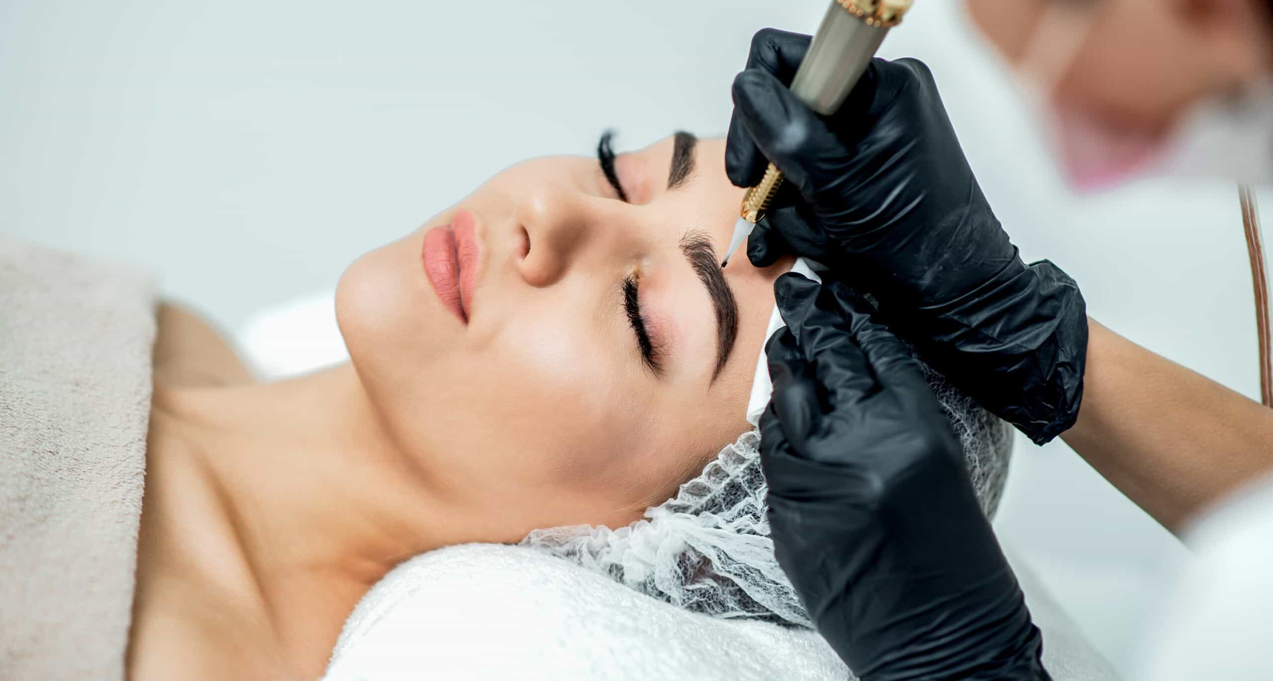 A woman is wrapped in a towel and wearing a hair net while she lays still getting her eyebrows tattooed.