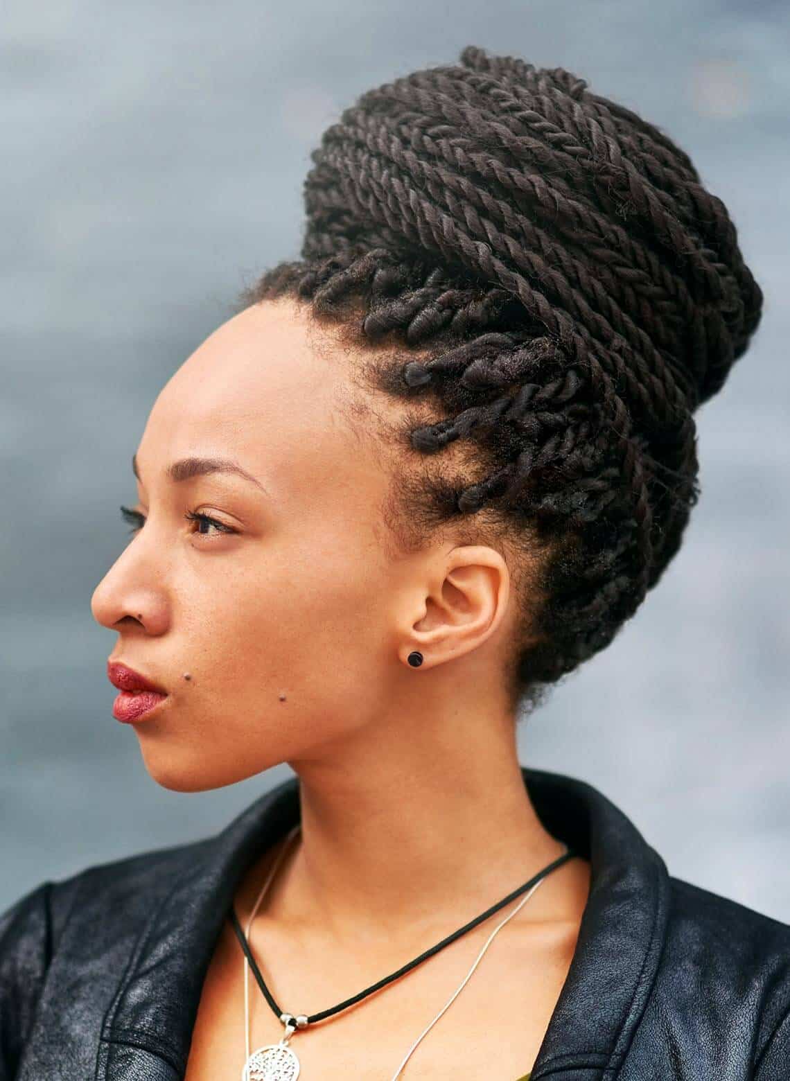 profile of person with Senegalese twists wrapped in a bun, wearing black stud earrings, a couple necklaces, a black leather jacket, and berry-colored lipstick