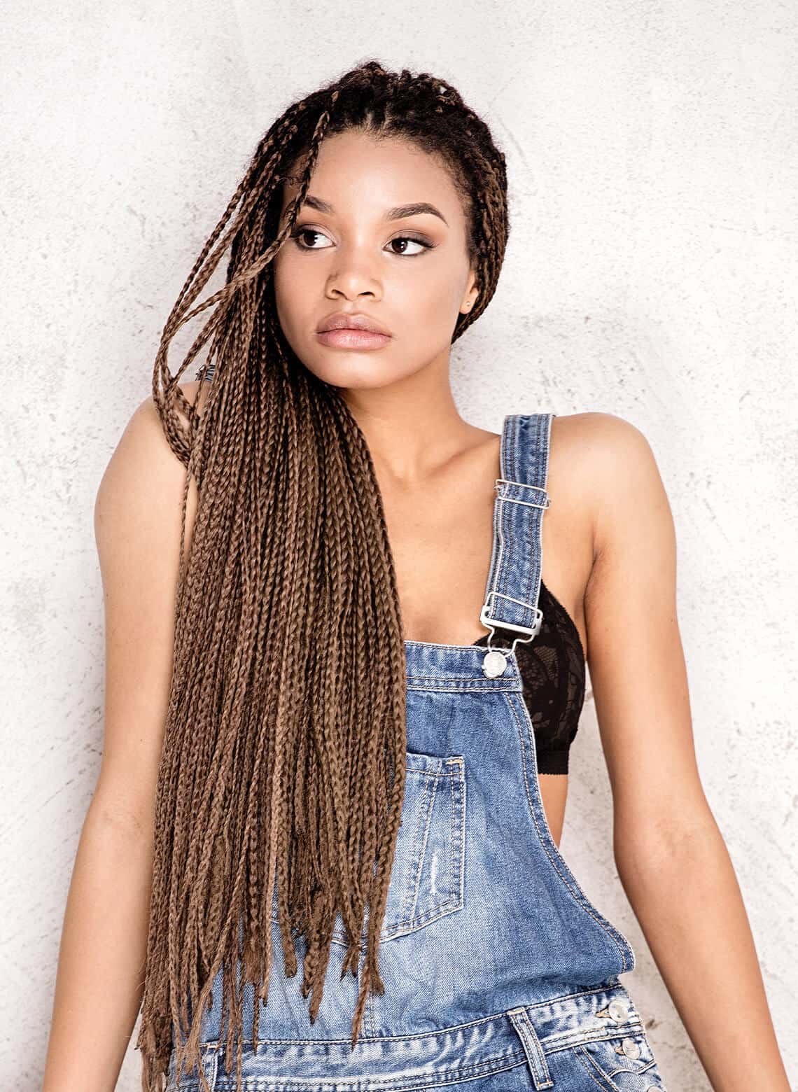 person with long microbraids swept over one shoulder wearing denim overalls and a black bralette underneath