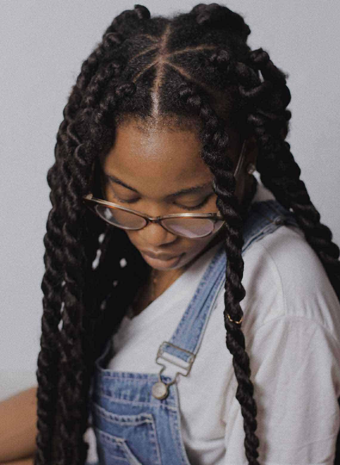 person looking down wearing long havana twists, glasses, white t-shirt and denim overalls