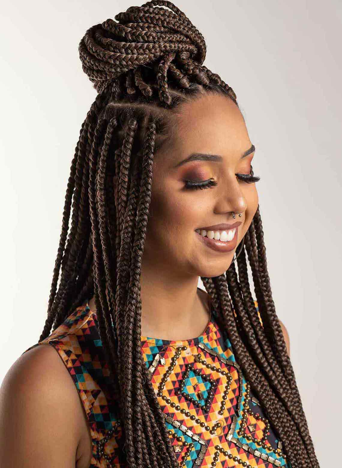 smiling person with eyes closed and wearing a braided bun with some braids hanging down on the sides, wearing a colorful geometric top and septum piercing