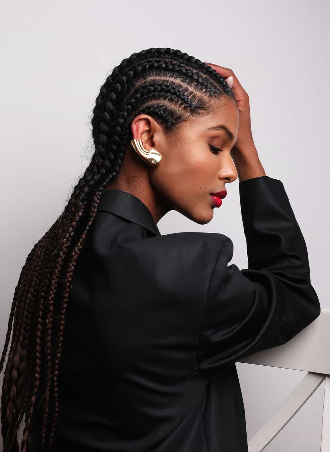 person with long stitch braids, gold ear cuff, and black blazer turned to the side and looking down