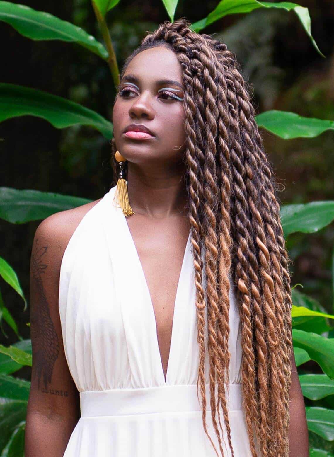 person with long brown and blonde marley twists looking the side, wearing long earrings and a white jumpsuit