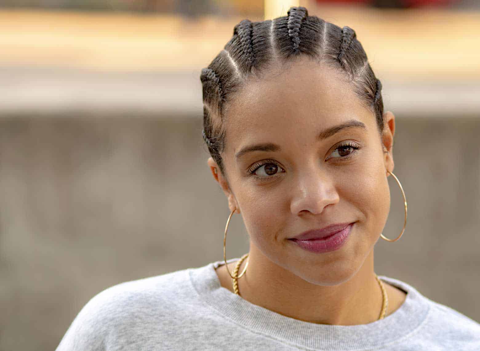 person with cornrows wearing a light gray sweater, big gold hoop earrings, and light berry-colored lipstick