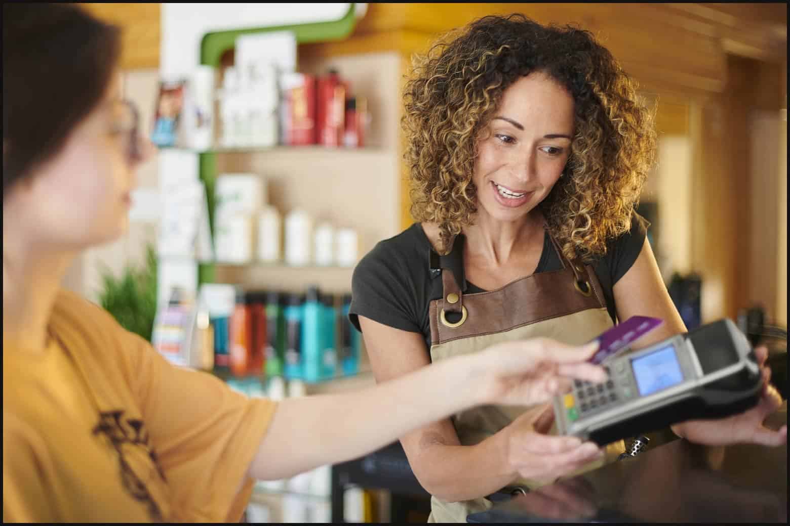 client wearing yellow shirt and glasses paying a salon owner with a credit card, salon owner wearing a black shirt and a smock is holding the card reader