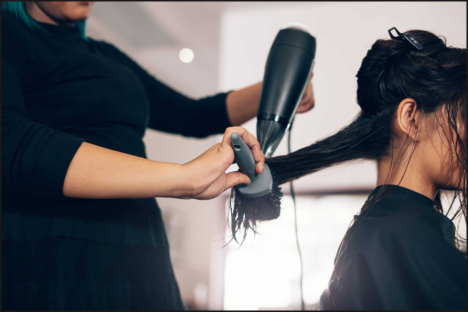 client wearing black smock with wet hair sitting in salon, close up on stylist blow-drying wet hair