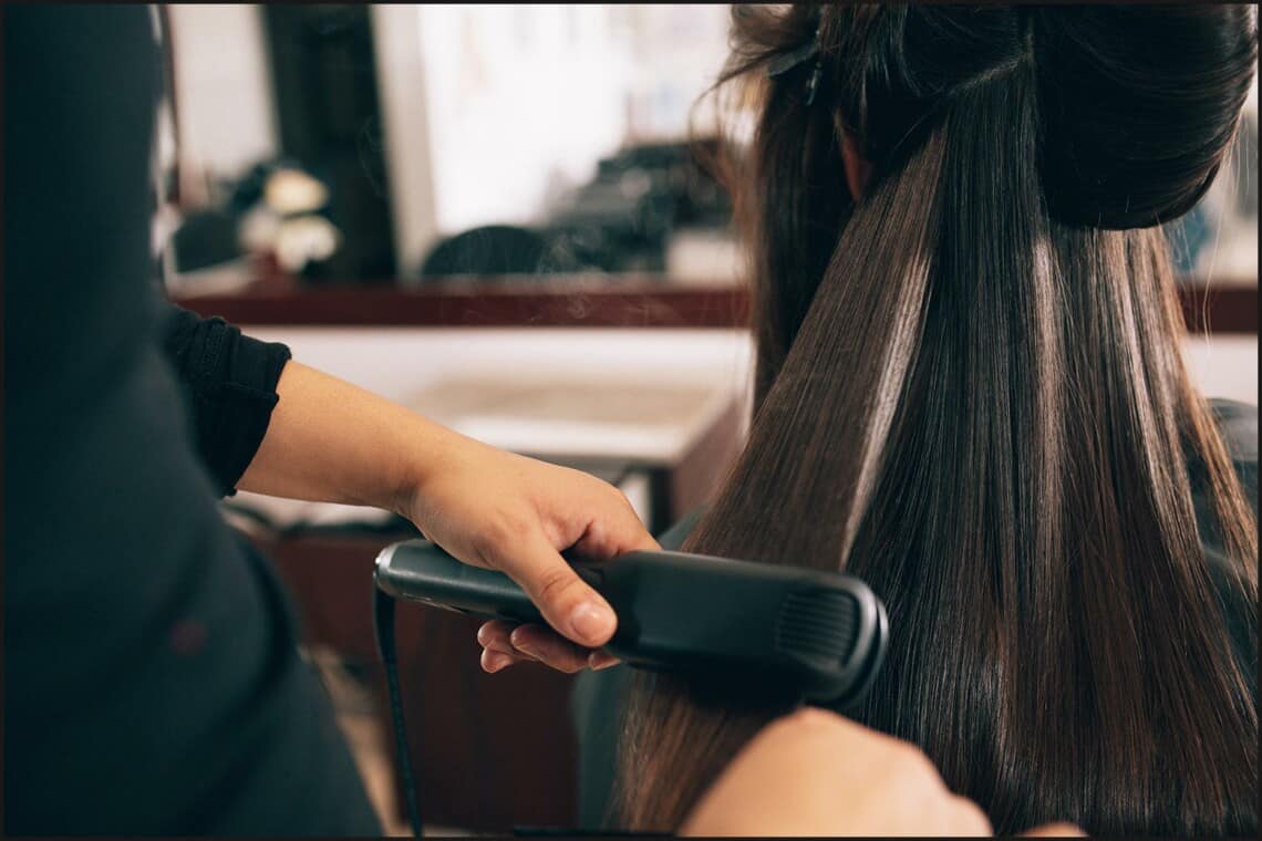 close up on stylist wearing black top flat ironing client’s brunette hair in a salon