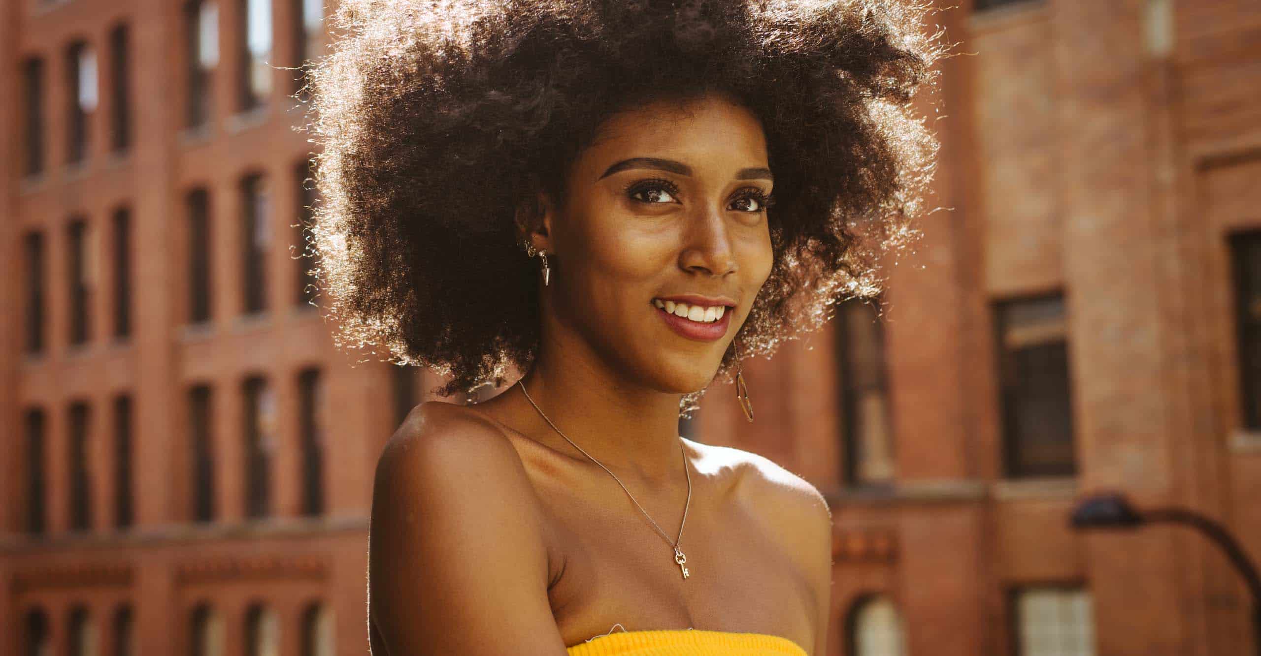 person with brunette natural hair walking in front a building, wearing a dainty necklace and yellow tube top