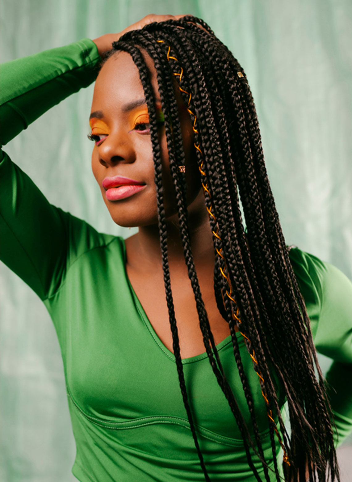 person wearing green top, orange eyeshadow, pink lipstick and accessorized box braids while looking down the side with one hand on the back of their head