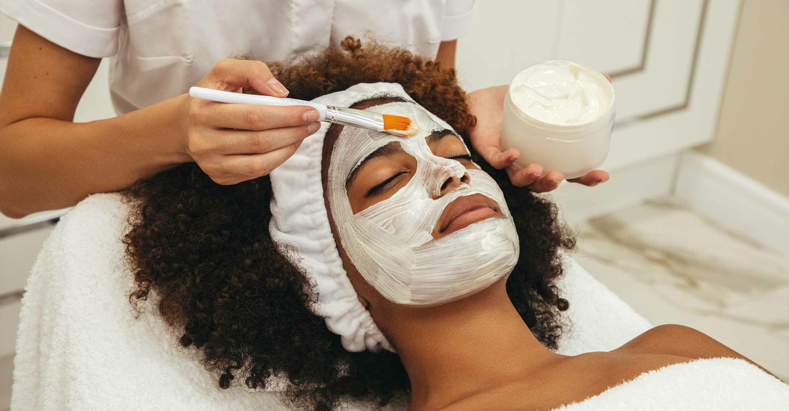 close-up on person with naturally coily hair, laying down and getting a facial applied in a salon