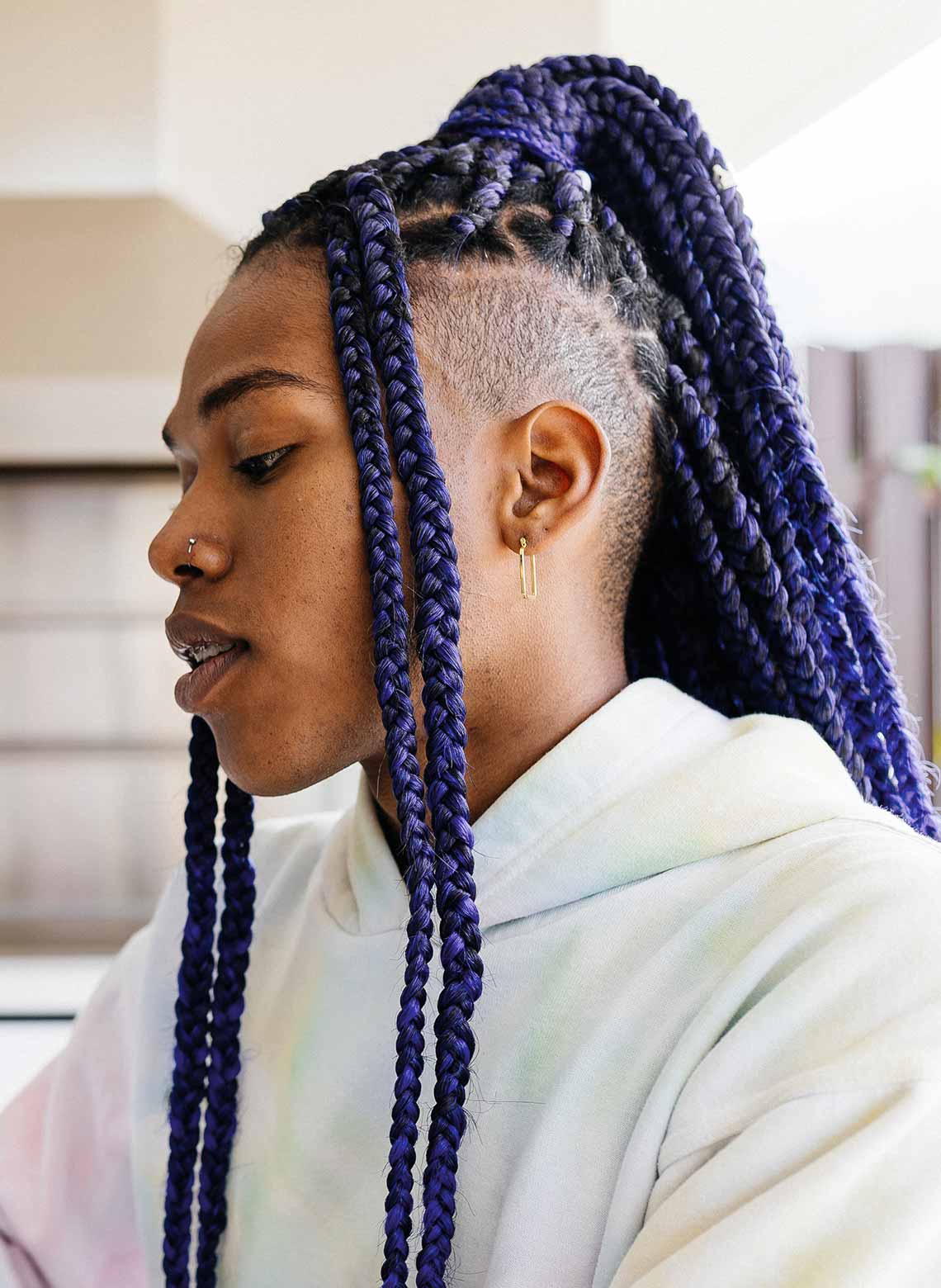 person wearing a light tie-dye sweater, nose ring, small hoop earrings, and purple knotless braids tied up in a ponytail, 