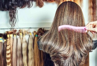 hand combing brunette wig with a pink comb in the foreground, collection of hair extensions of different hair colors hung along the wall in the background