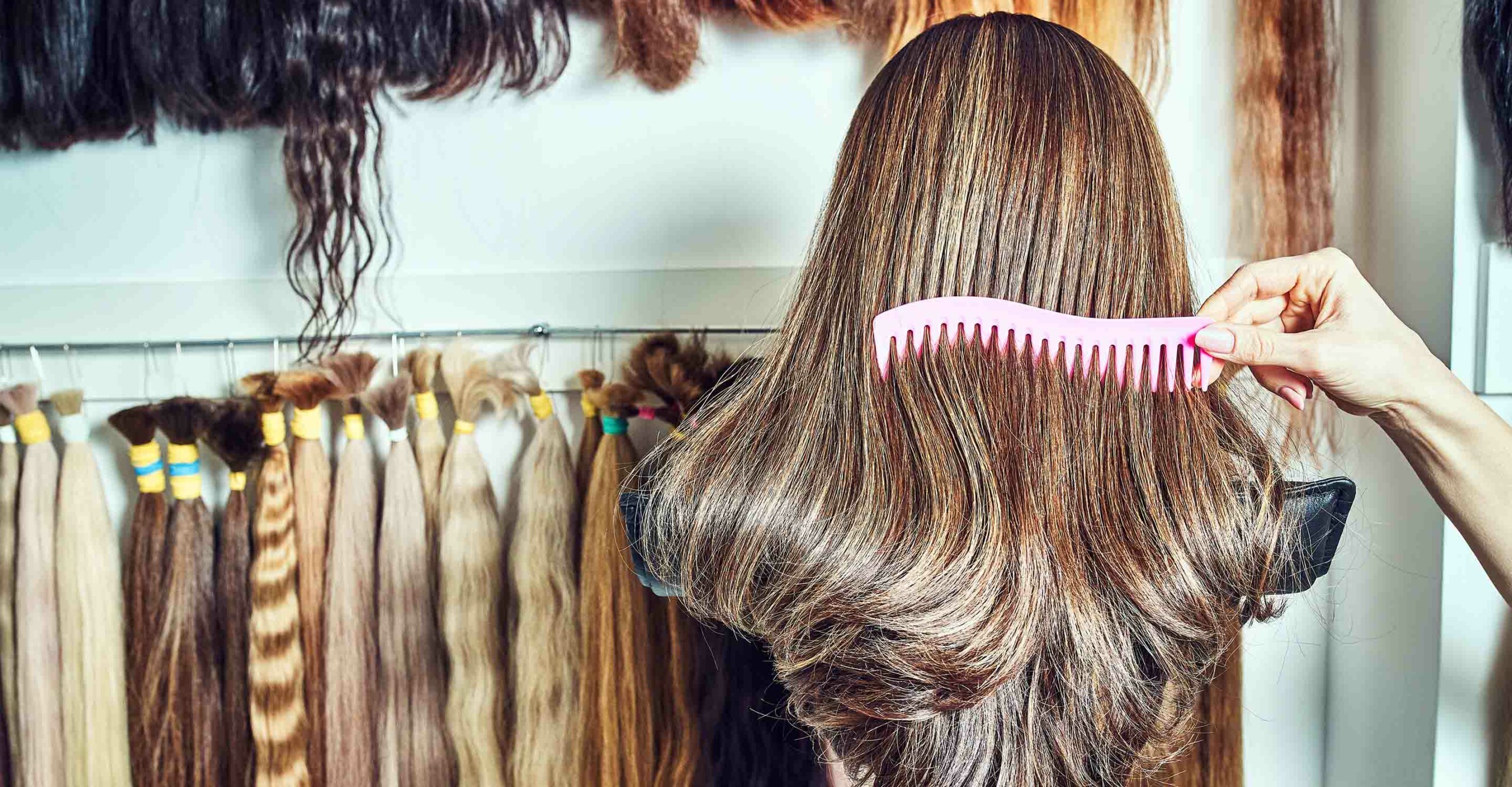 hand combing brunette wig with a pink comb in the foreground, collection of hair extensions of different hair colors hung along the wall in the background
