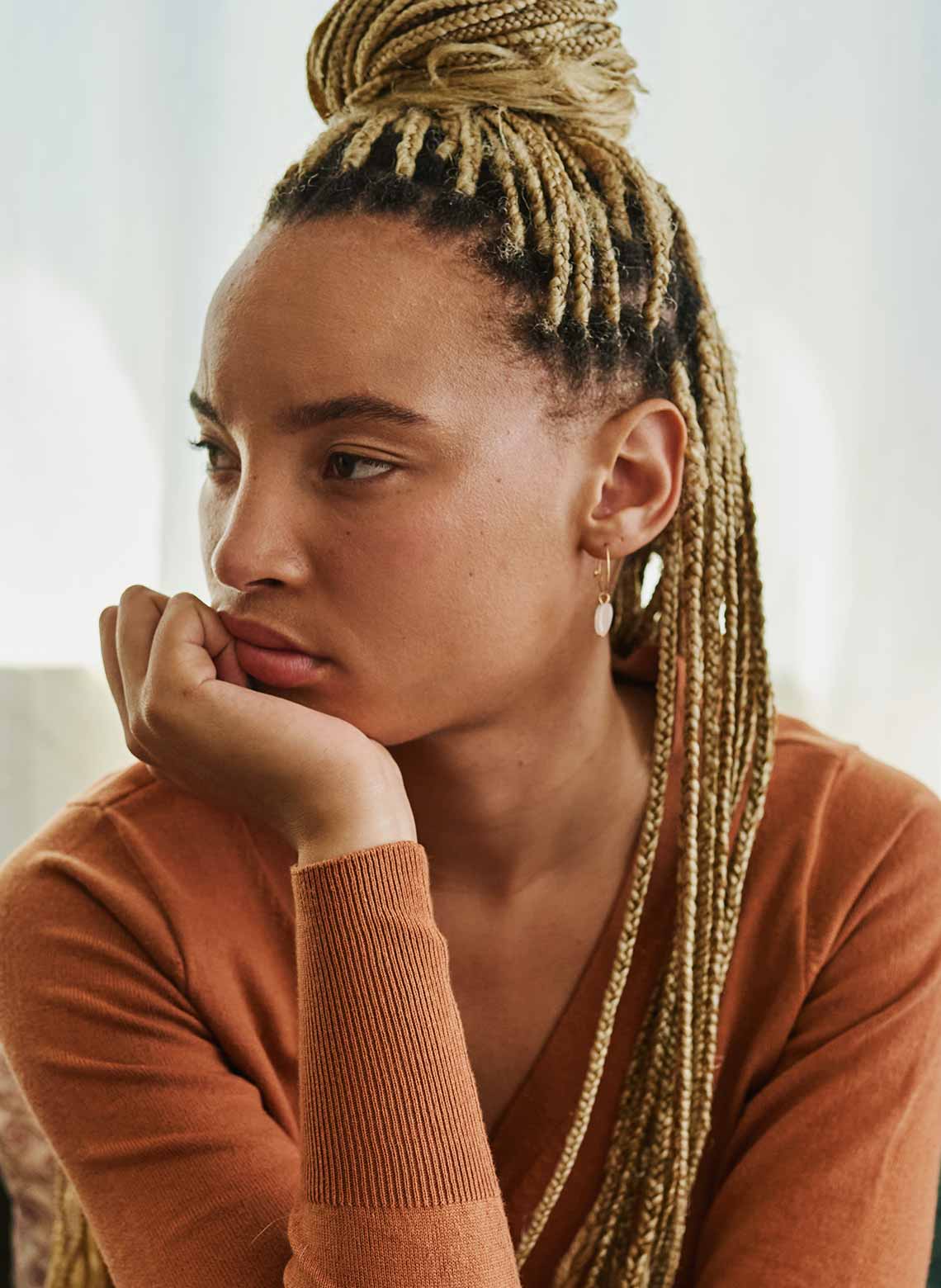 person wearing burnt orange long-sleeve shirt, half-up, half-down blonde knotless braids, with hand on their chin and looking to the side
