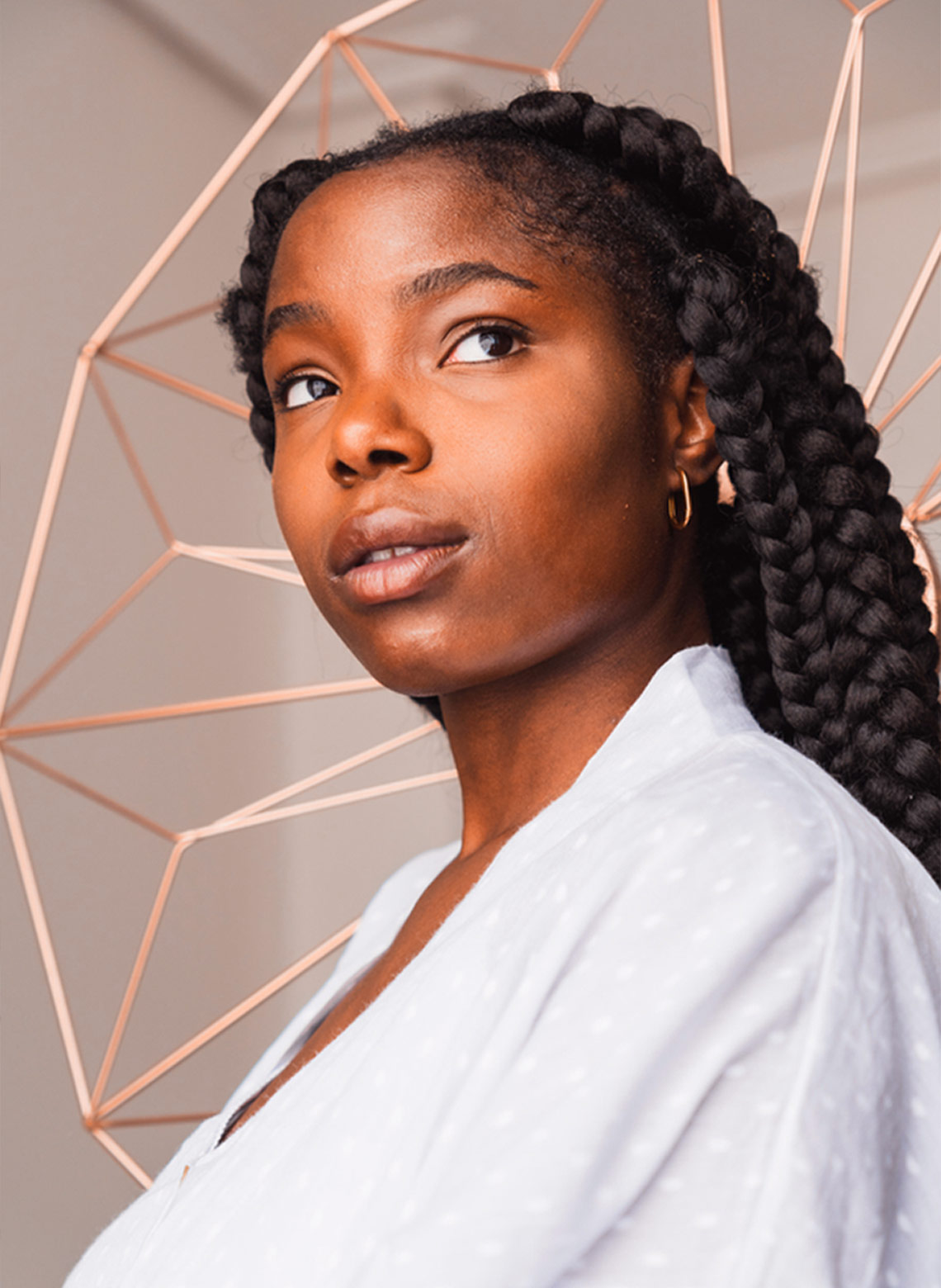 person wearing white blouse with subtle polka dot details, hoop earrings, and jumbo box braids while standing in front of a rose gold geometric background