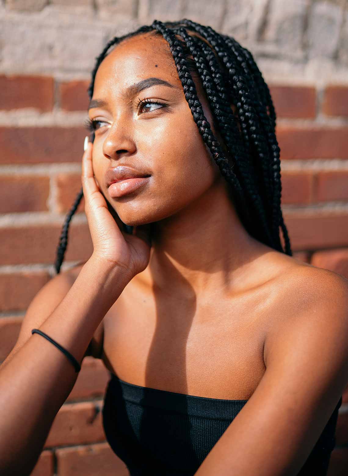 person wearing a black tube top, black hair tie around their arm, long medium box braids, and their hand on their face while looking to the side
