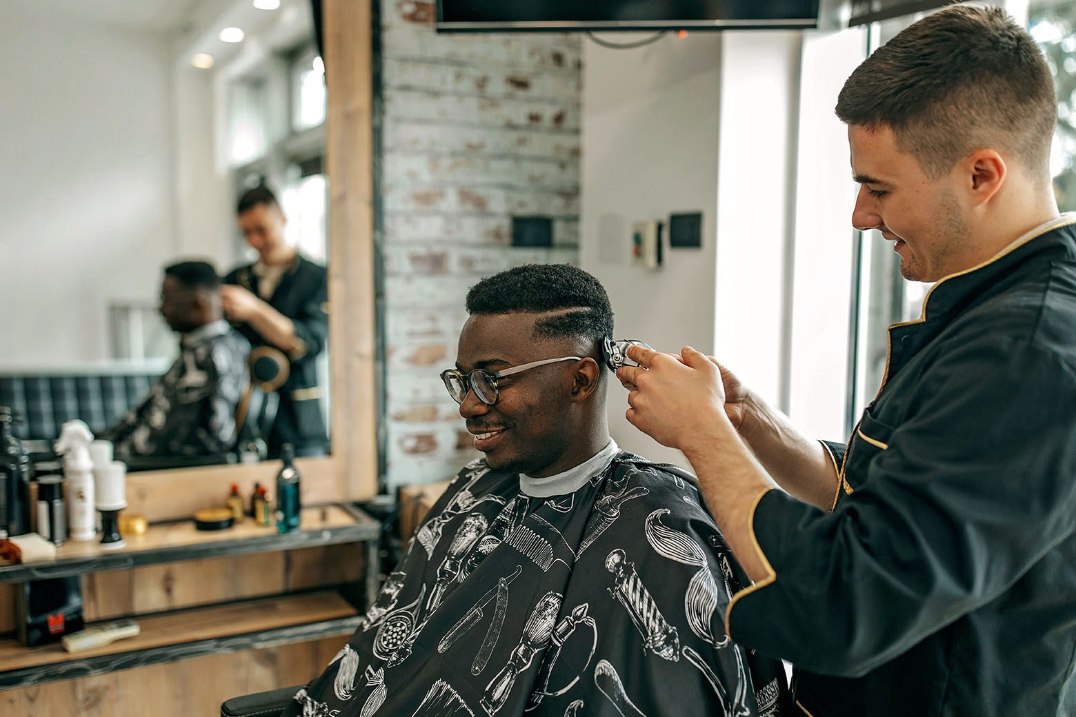 A person sitting in the barber chair, wearing a black smock with white barbershop illustrations, with barber cutting cutting hair