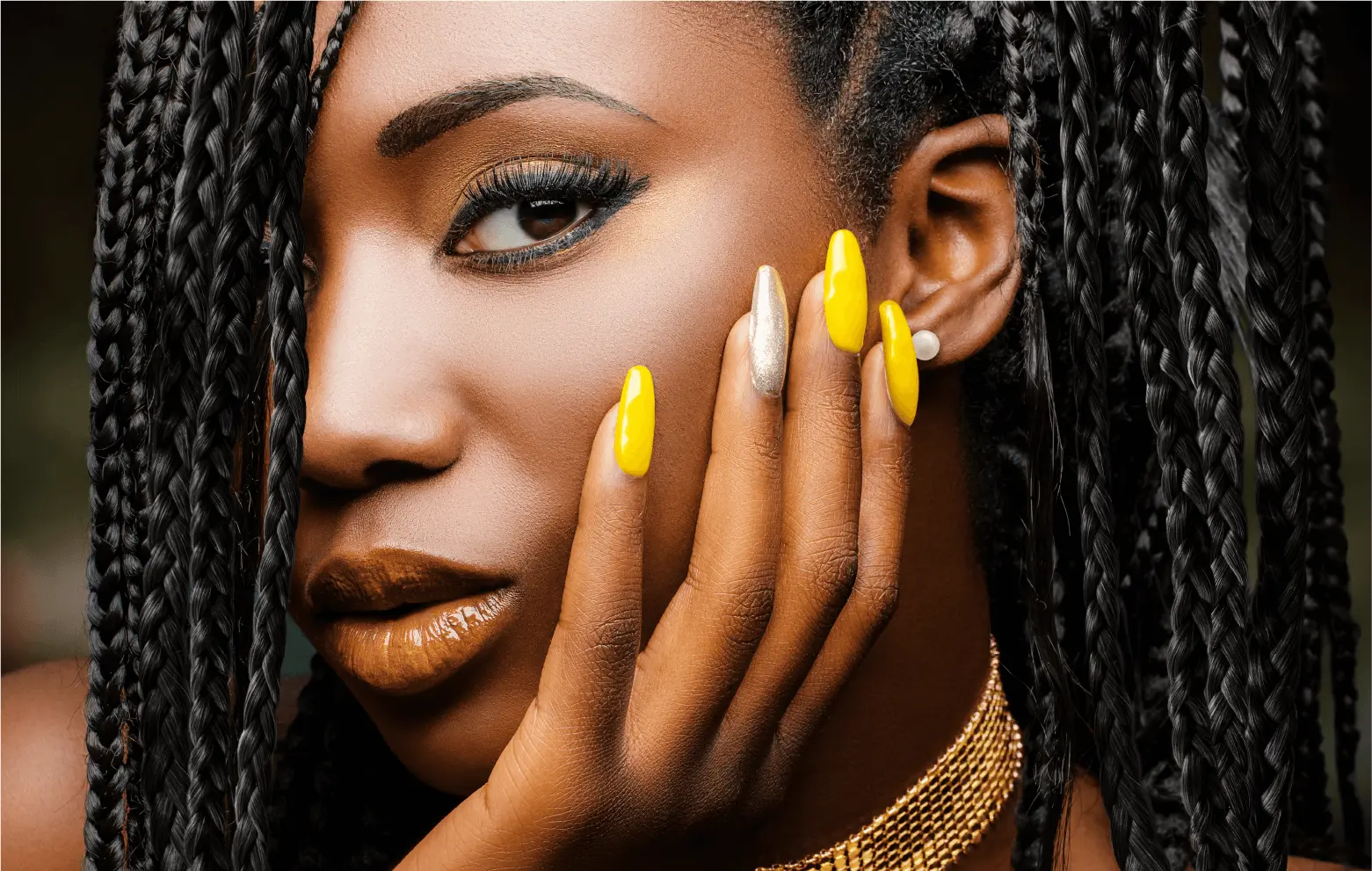 close-up of person with long braids, full glam makeup, and hand with nail art touching the side of their face