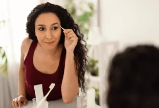 woman shaping and tweezing her eyebrows while looking in a mirror